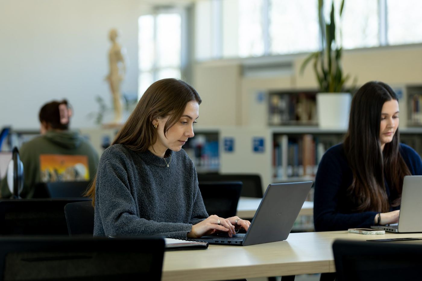 A student uses a laptop at a library desk with bookshelves and other students in the background.