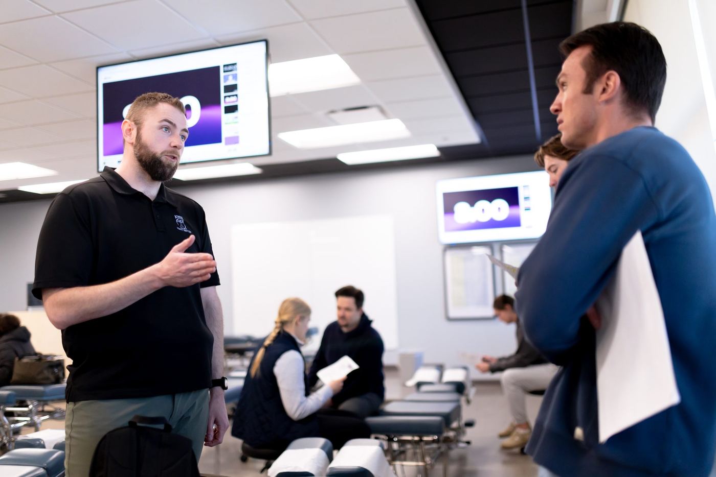 Instructor in black polo speaks to students in a classroom with examination tables and screens showing a timer.