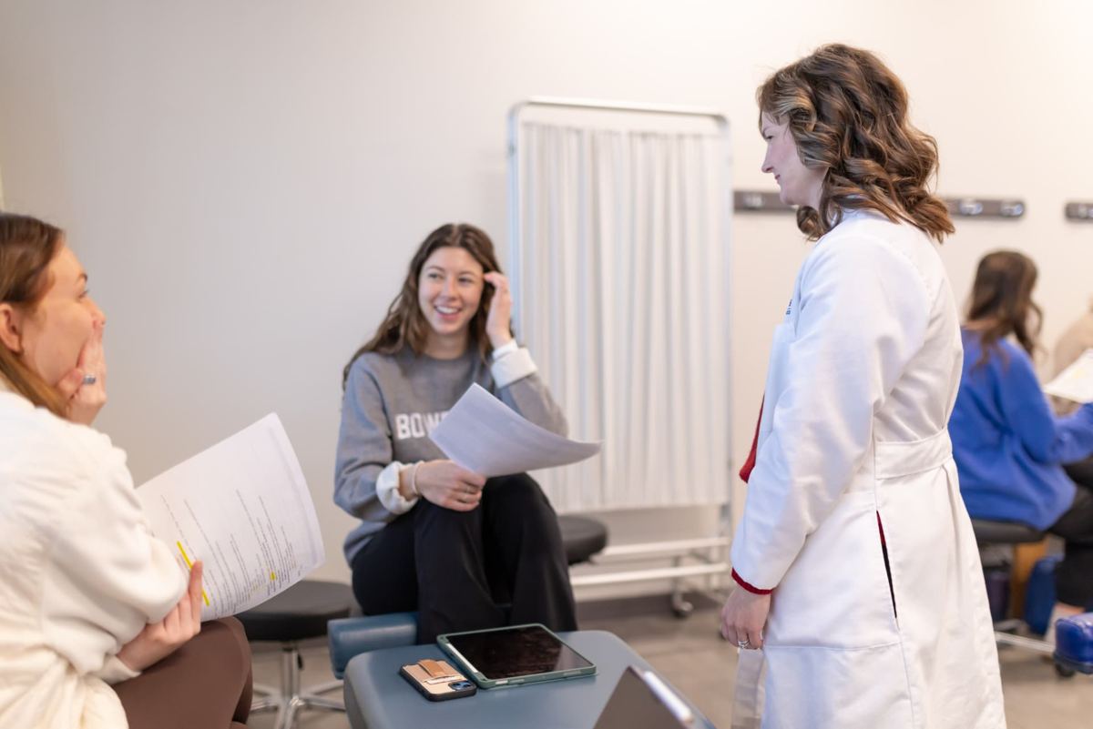 A Logan University clinician in a white coat talking with smiling students in a clinical skills room