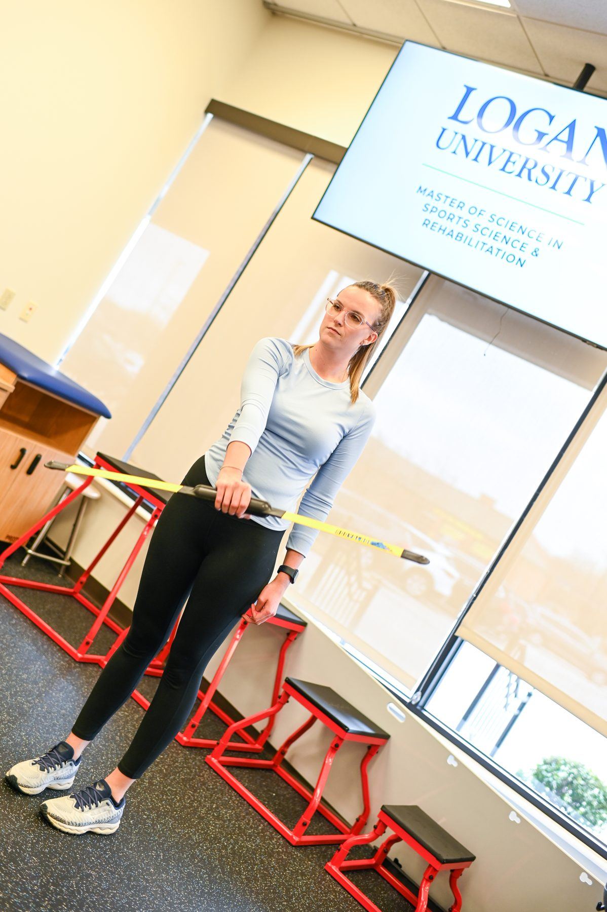 Female student in glasses and light blue shirt holds a yellow exercise bar in a rehab lab setting.