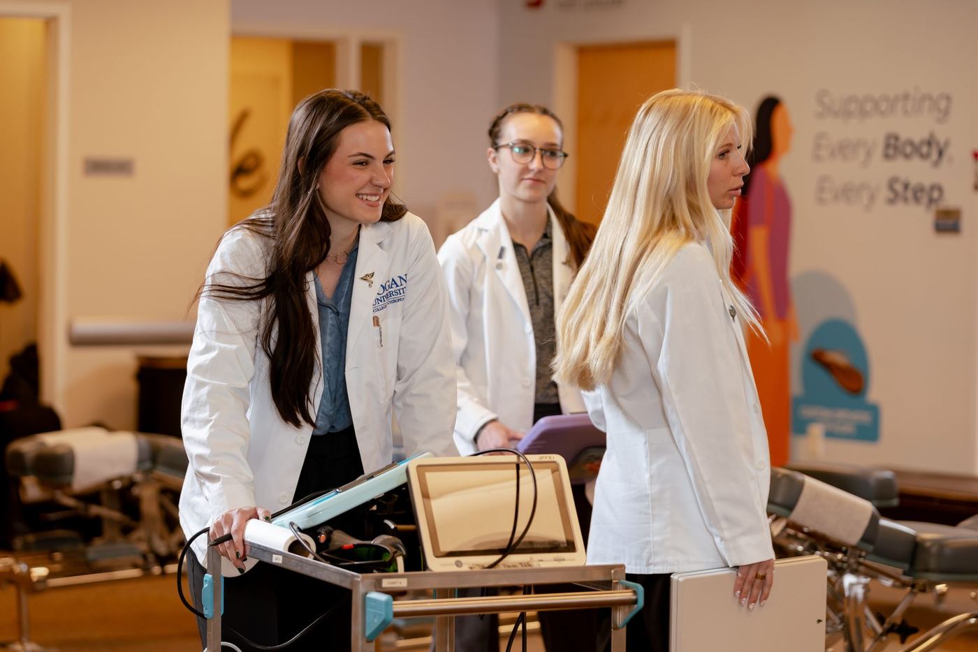 Three women in white lab coats collaborate in a lab with medical equipment and a branded wall.