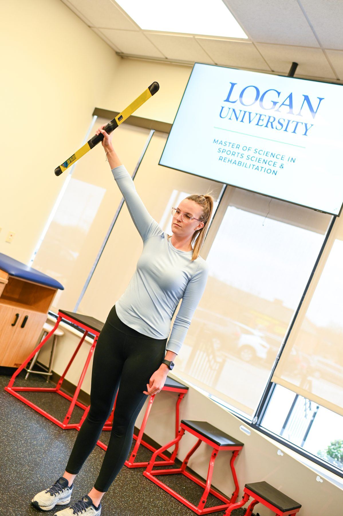 Student in athletic wear holds yellow rehab bar overhead, Logan University logo on screen in background.
