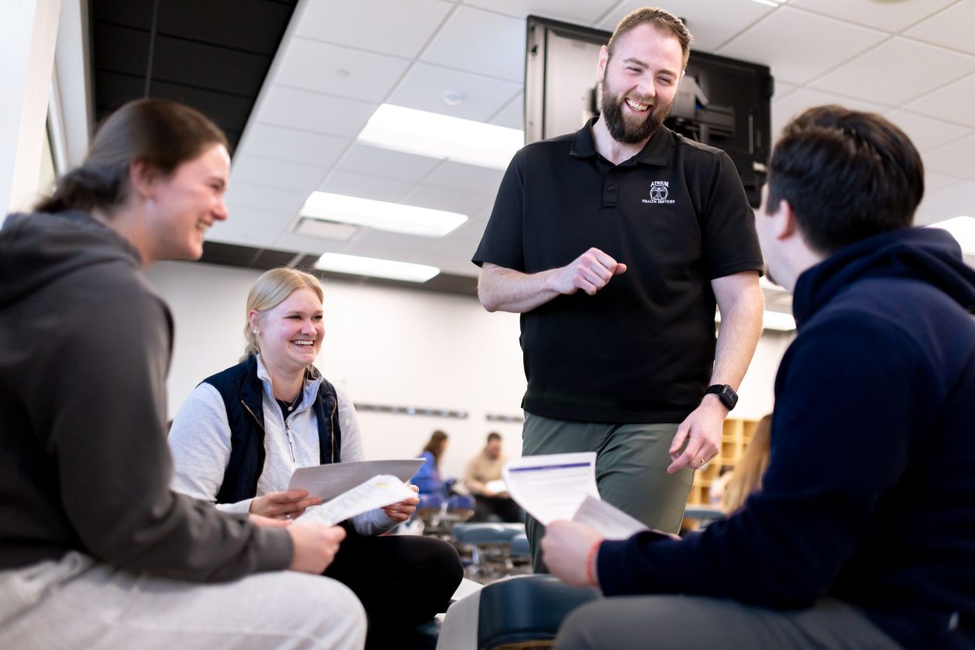 Instructor in black polo converses with three students holding papers in a modern classroom.