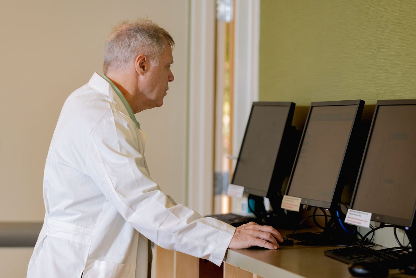 Man in a white lab coat intently viewing multiple computer monitors at a desk.