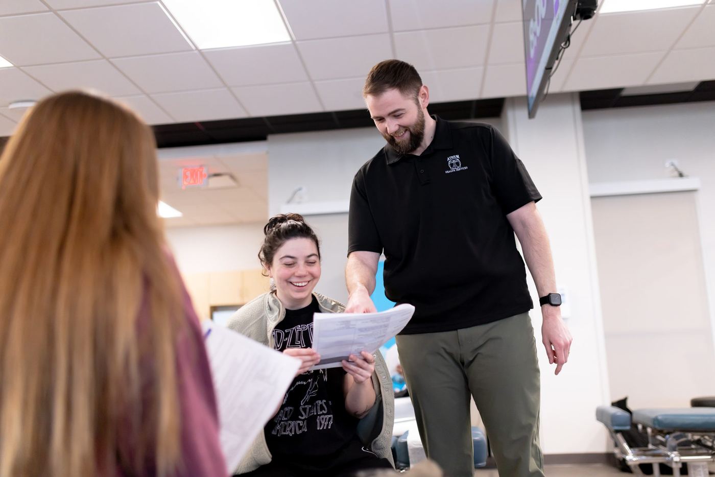 Smiling man in black polo helping female student review papers, another student in foreground.