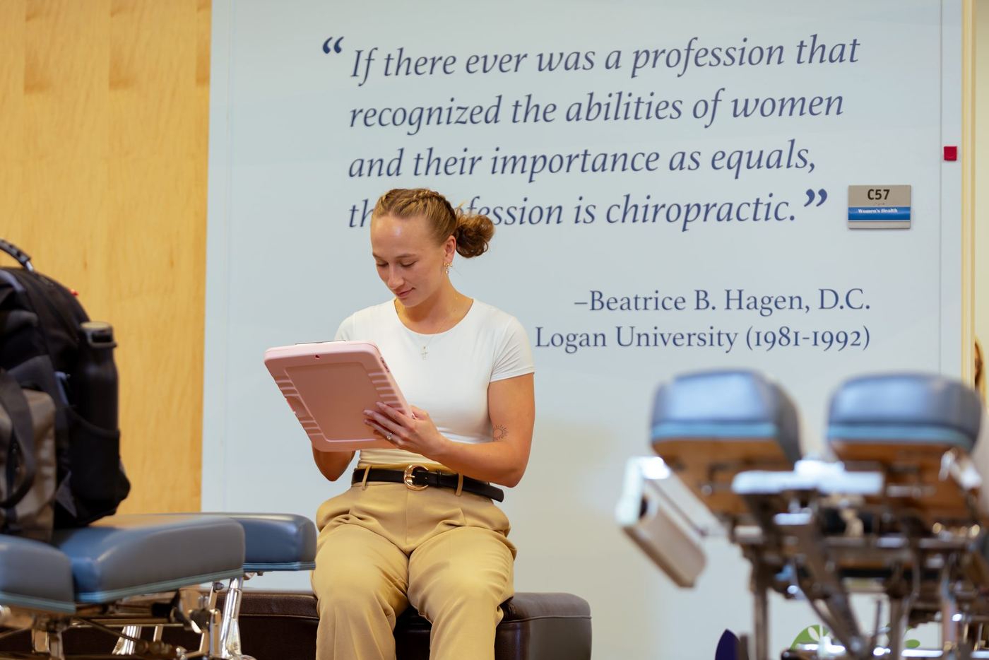 Young woman in clinic setting uses a pink tablet, chiropractic tables and quote visible in background.