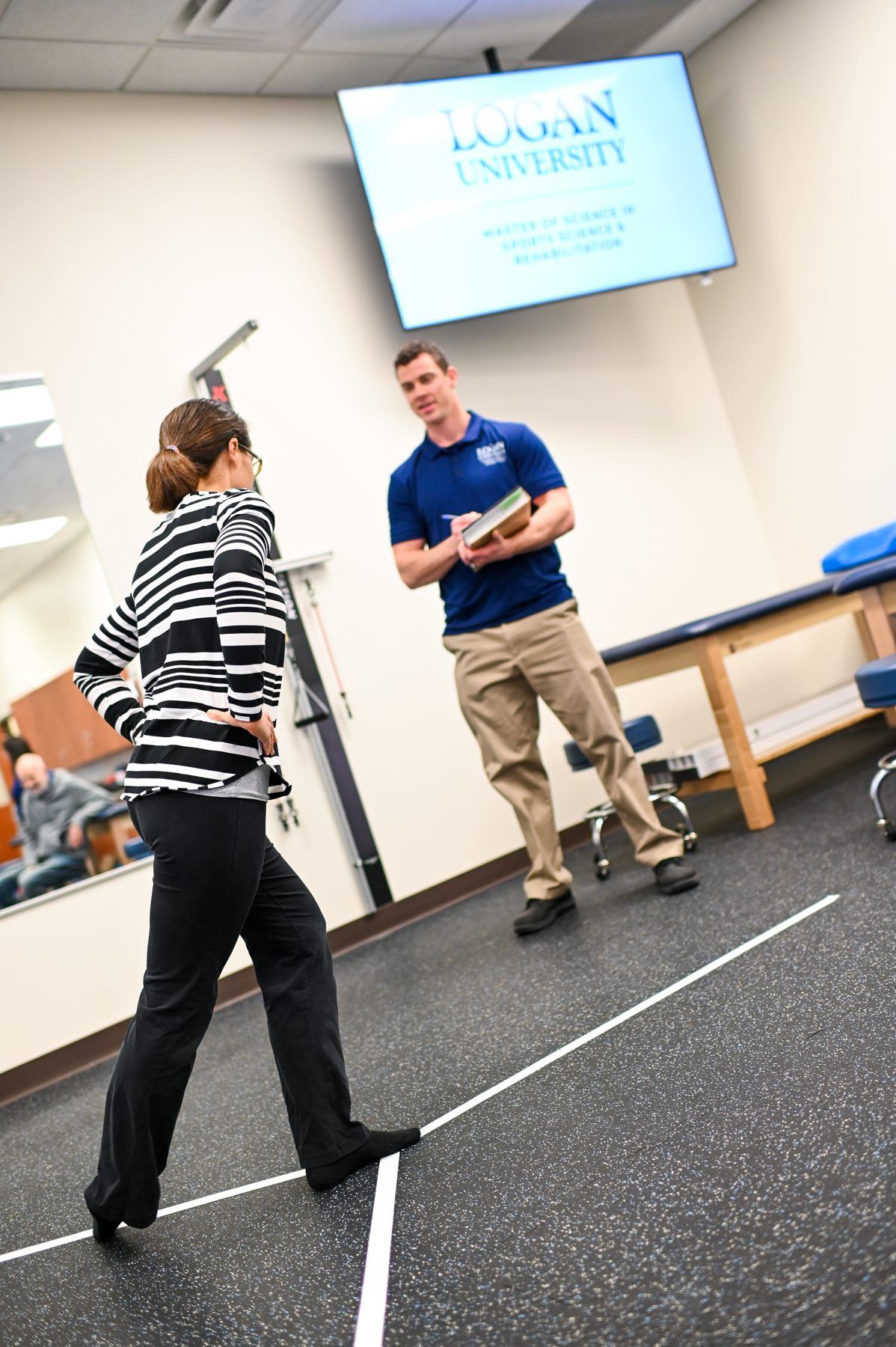 Physical therapy student doing balance exercise on marked floor, instructor observing and taking notes.