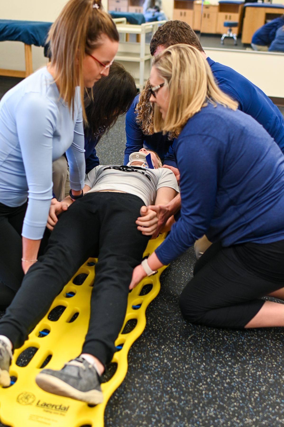 Four students stabilize a prone person on a yellow spine board during a medical training exercise.