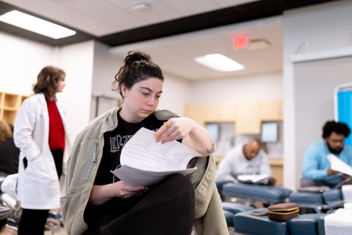 Young woman in black t-shirt and green fleece reviews papers in a brightly lit classroom setting.