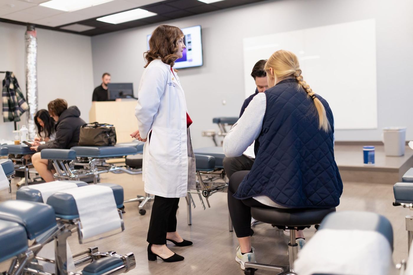 Instructor in a white lab coat teaches students at chiropractic adjustment tables in a classroom.