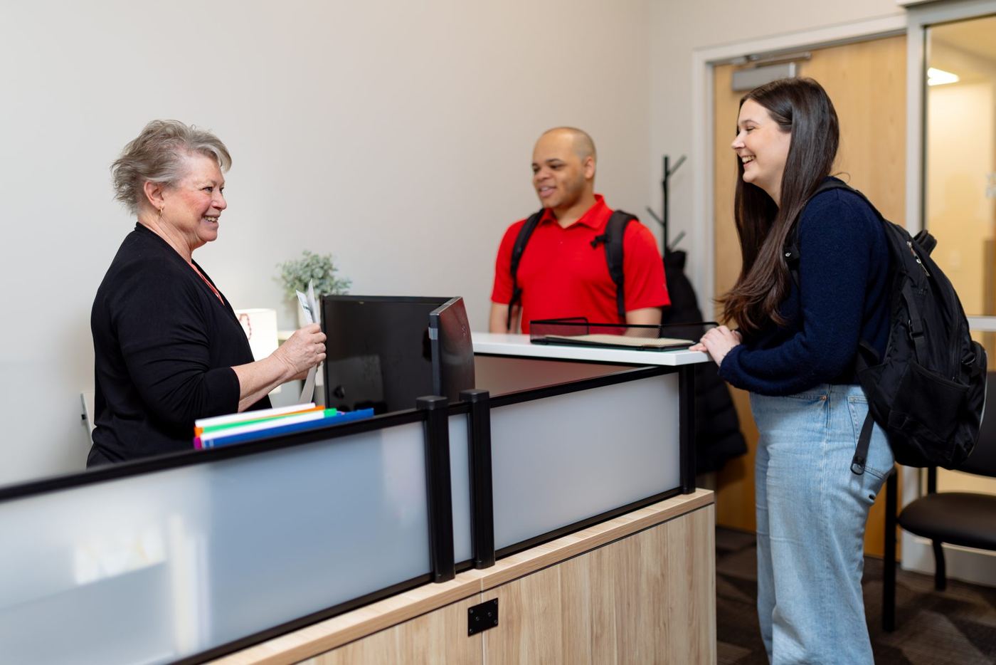 Smiling older woman at reception desk assists a young female student, a male student stands nearby.
