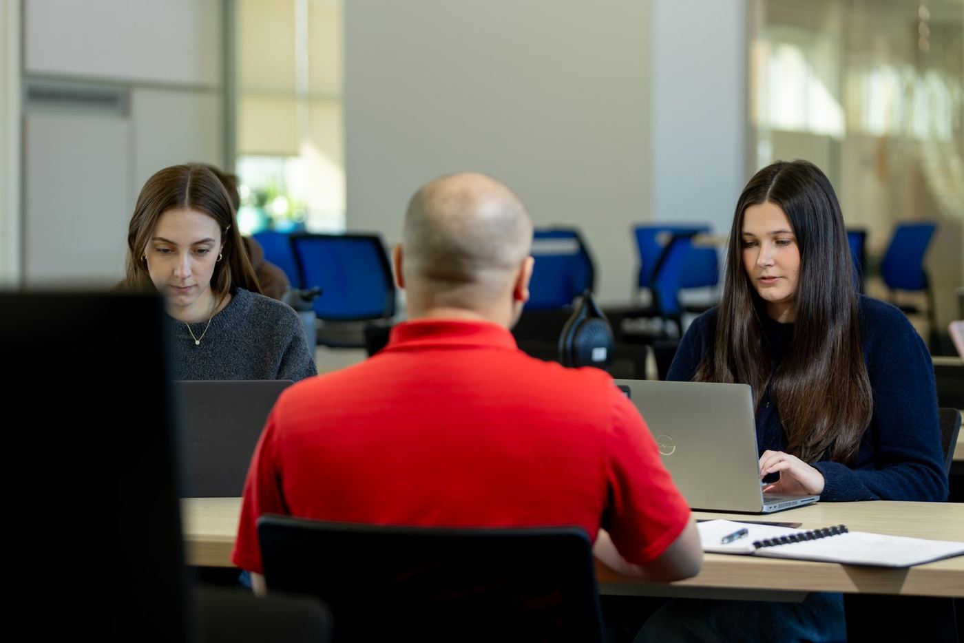 Three students work on laptops at modern desks in a bright classroom.