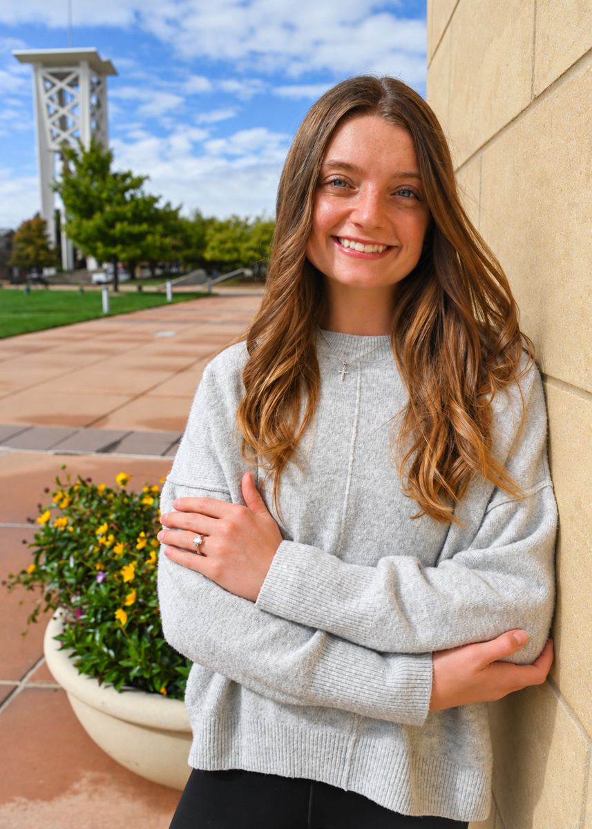 Logan University student Jaiden Root smiling with arms crossed, leaning on a campus wall with the bell tower behind her