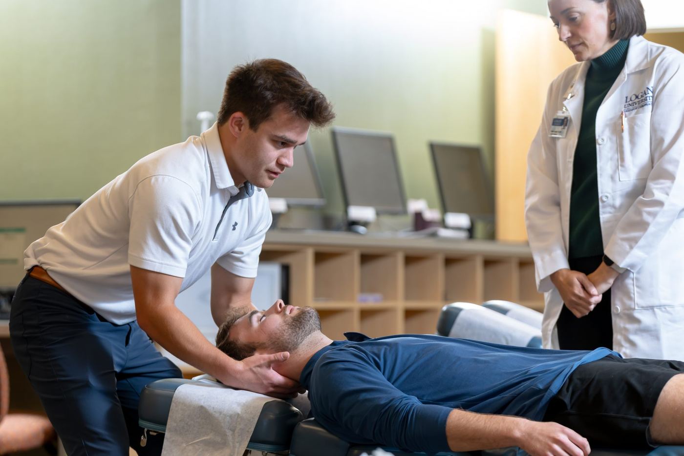 Logan student performs neck adjustment on patient as female instructor in lab coat observes.