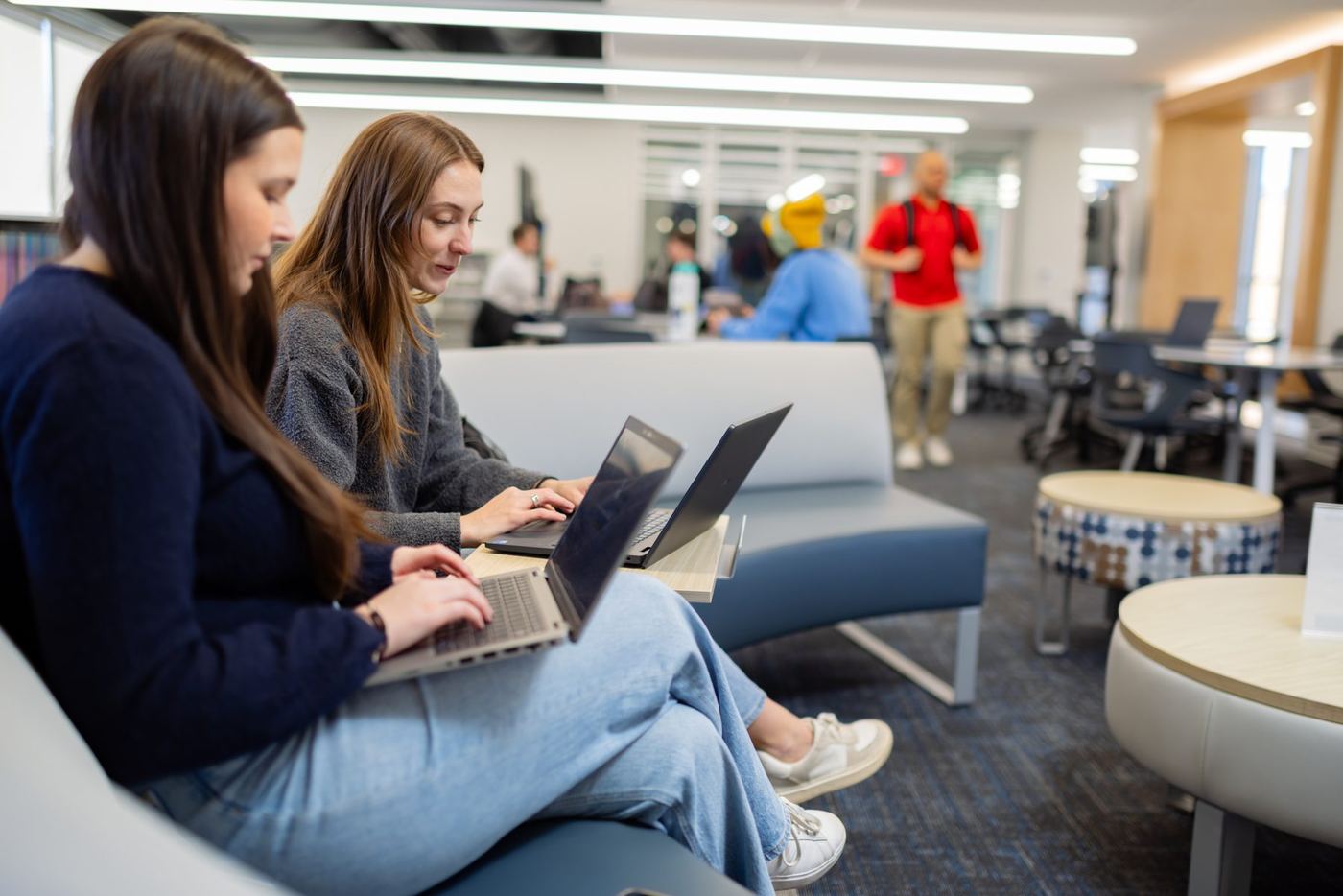 Two students on a blue bench focused on laptops in a bright, modern university study area.