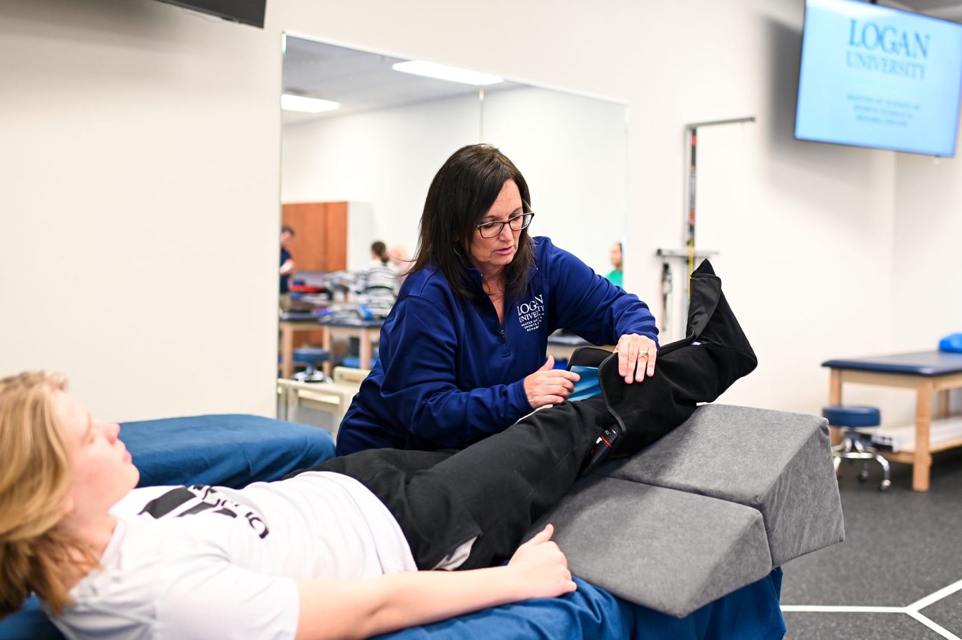 Woman in Logan jacket assists patient on examination table with leg compression device in clinic.
