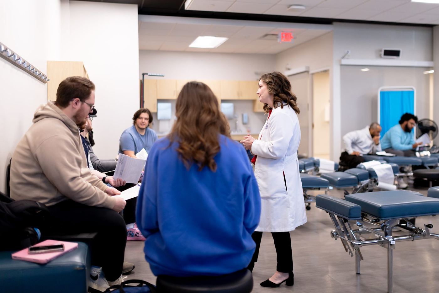 PA students in a modern medical lab listening to an instructor, surrounded by adjustment tables.