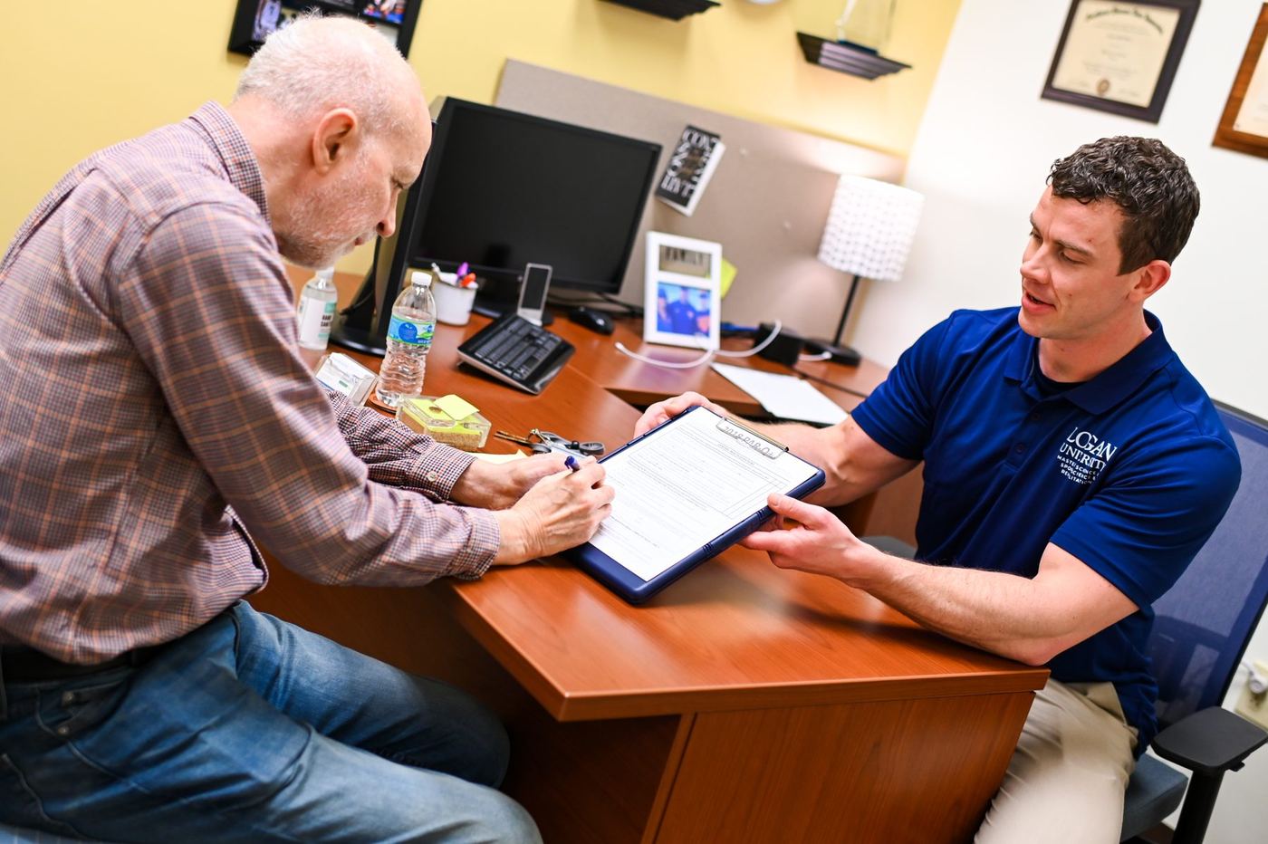 Older man signs document on clipboard held by younger man in Logan University polo shirt across a desk.