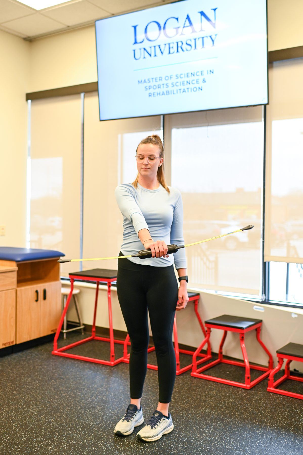 Woman in sports science lab holds flexible bar, screen shows 'Logan University: MS Sports Science'.
