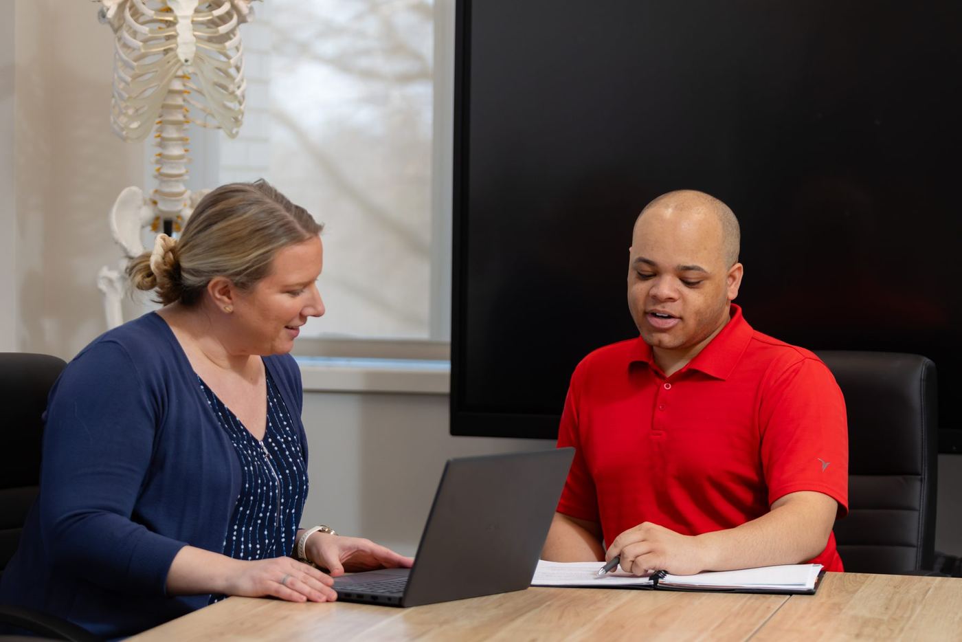 Two students collaborating at a table with a laptop, documents, and a skeleton model in background.