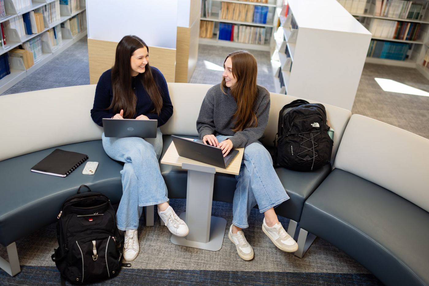 Two students work on laptops and chat in a bright, modern library area at Logan University.