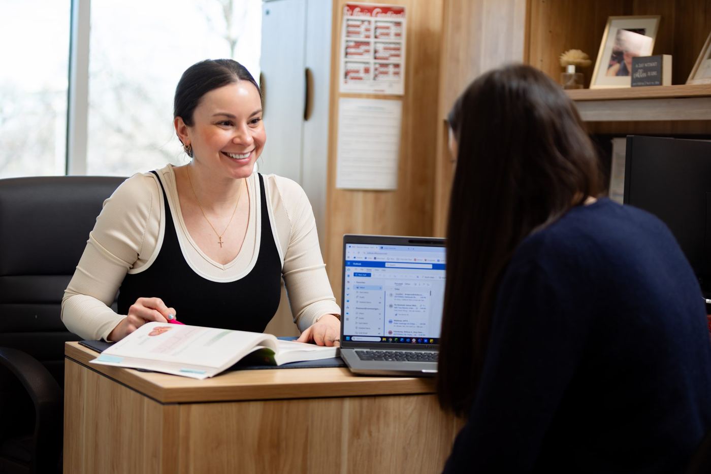 Smiling woman at a desk with a laptop and open book, looking at another person.
