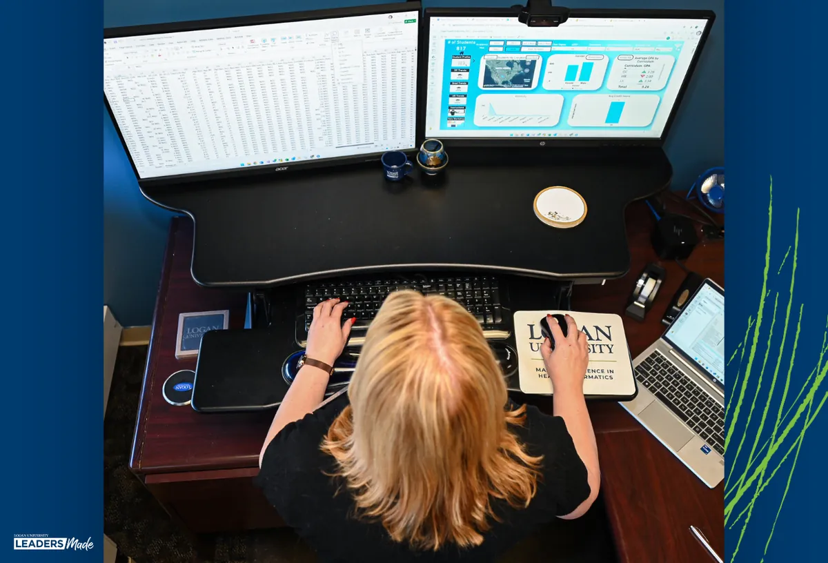 A person works at a dual-monitor computer setup analyzing data spreadsheets and charts, with a Logan University Master of Science in Health Informatics mousepad visible on the desk.