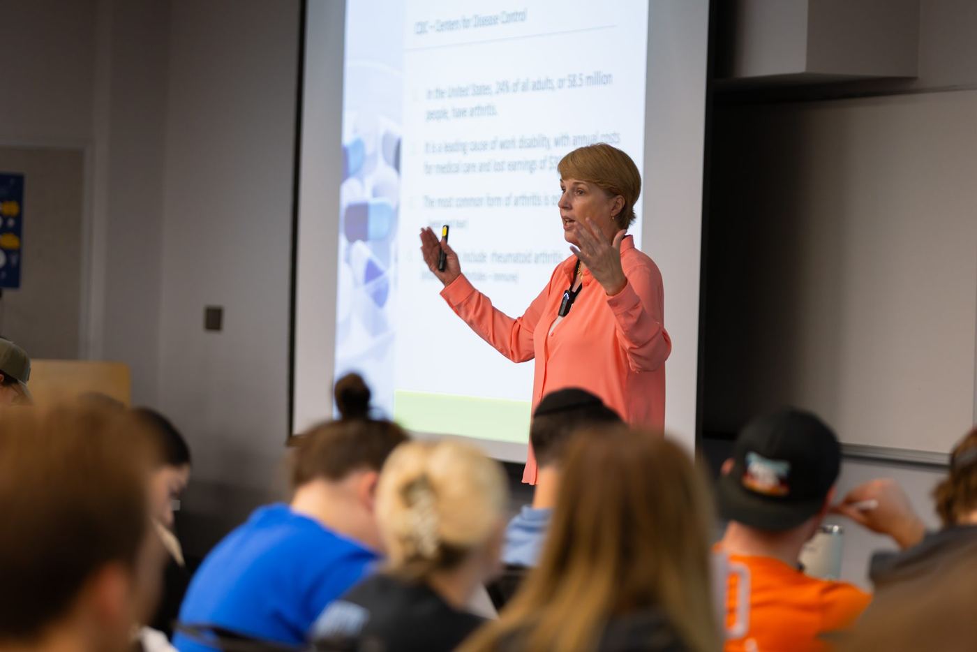 Presenter in orange shirt gestures, speaking to students in a classroom with an arthritis presentation on screen.