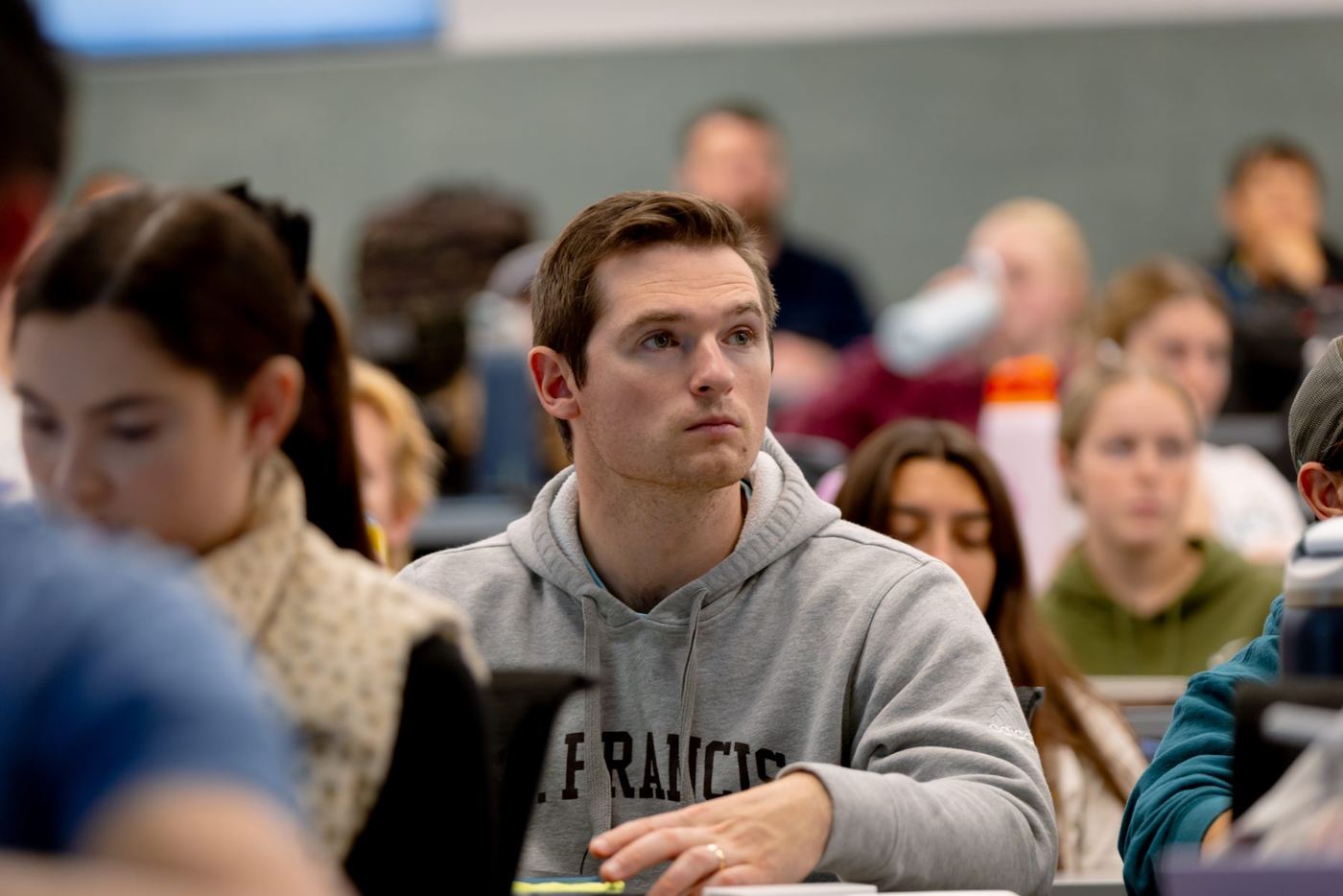 Male student in gray hoodie with 'FRANCIS' listens intently in a classroom with other students.