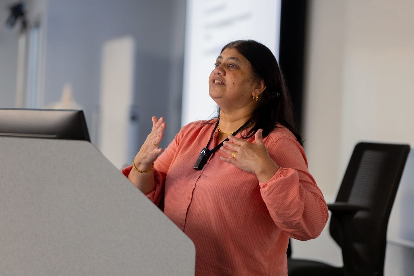 A woman in a peach blouse speaks animatedly at a lectern in a modern conference room.