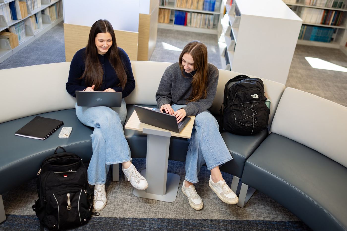 Two female students smile while working on laptops on a curved bench in a modern library.