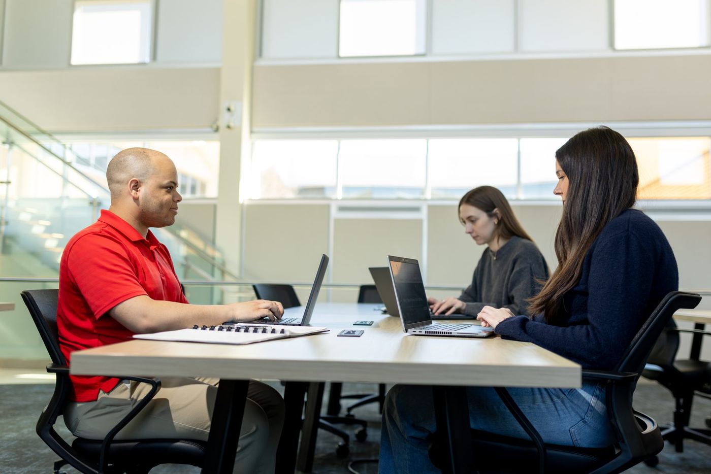 Three students work on laptops at a table in a bright, modern university building with large windows.
