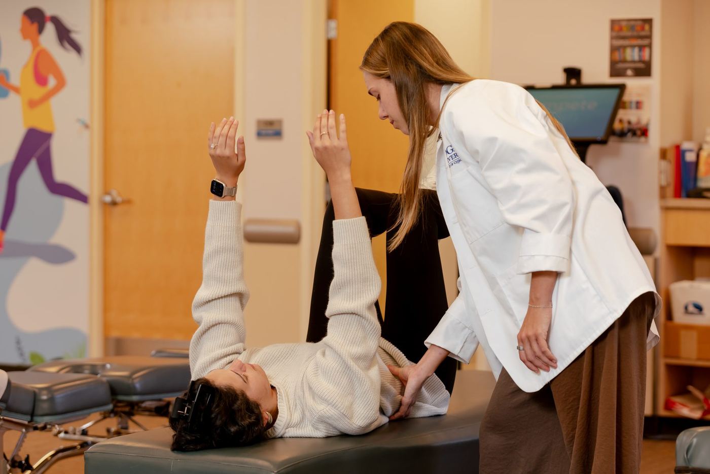 Logan DC student or doctor in white coat examines a patient lying on a chiropractic table with legs and arms raised.