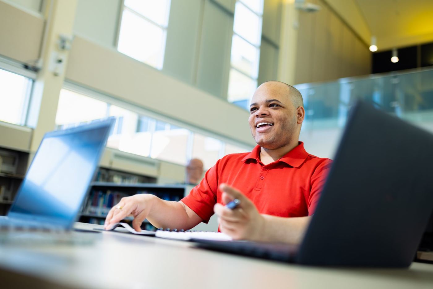 Smiling student in red polo works at desk with two laptops and notebook in a modern library.