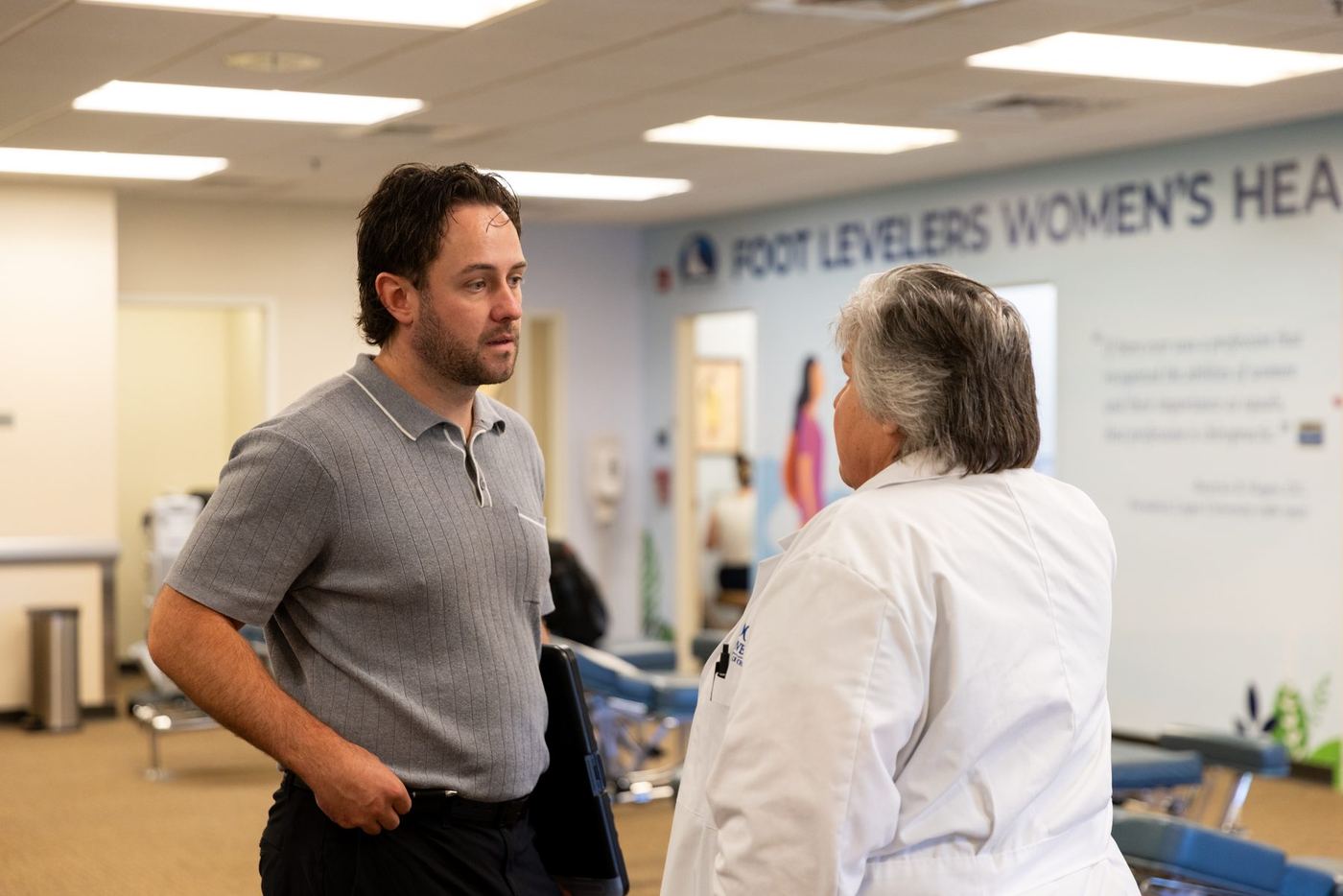 A man in a gray polo talks to a woman in a white lab coat in a brightly lit facility.