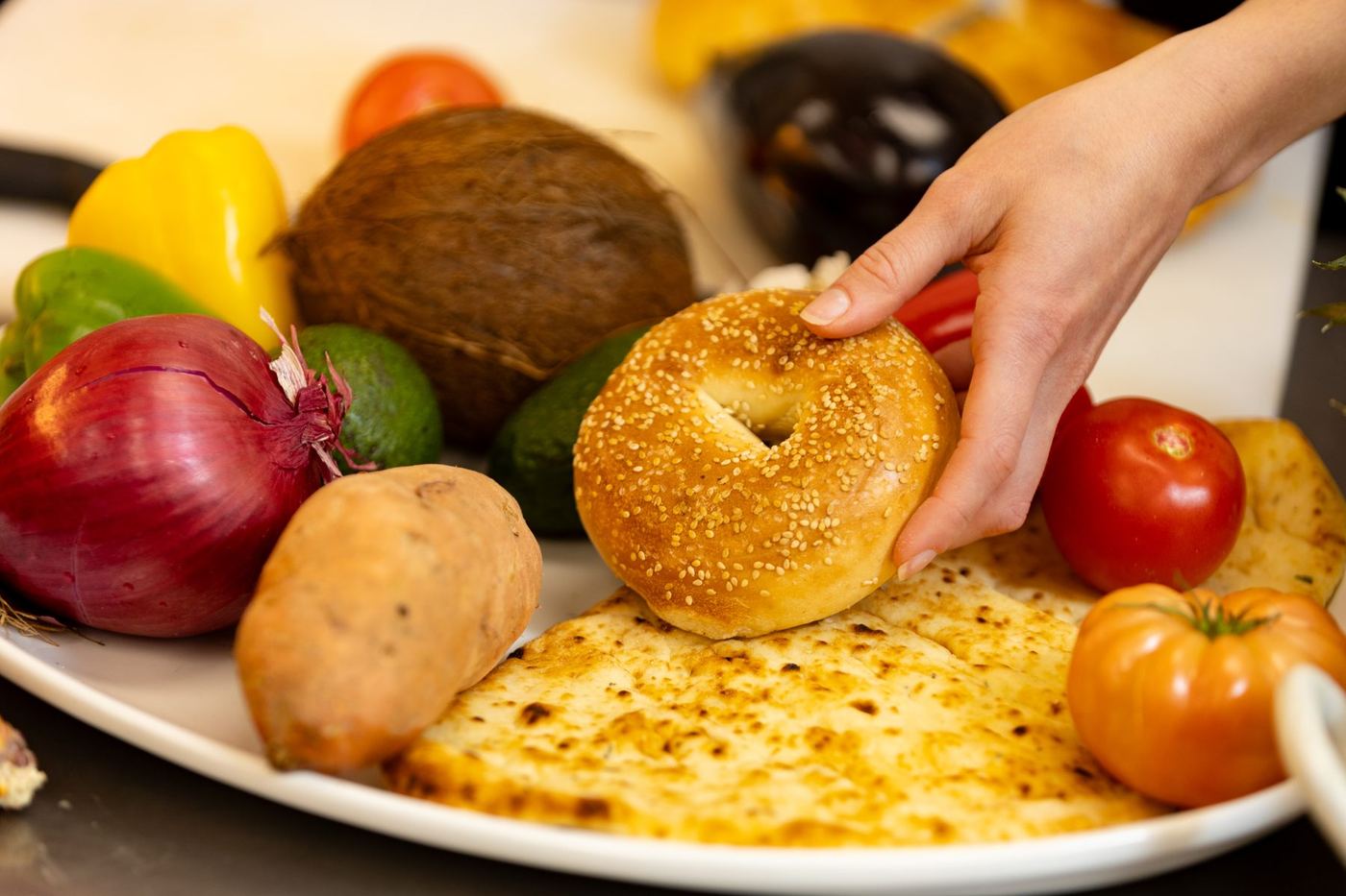 Hand reaching for a sesame bagel on a white platter with various vegetables like bell peppers, tomatoes, avocado.