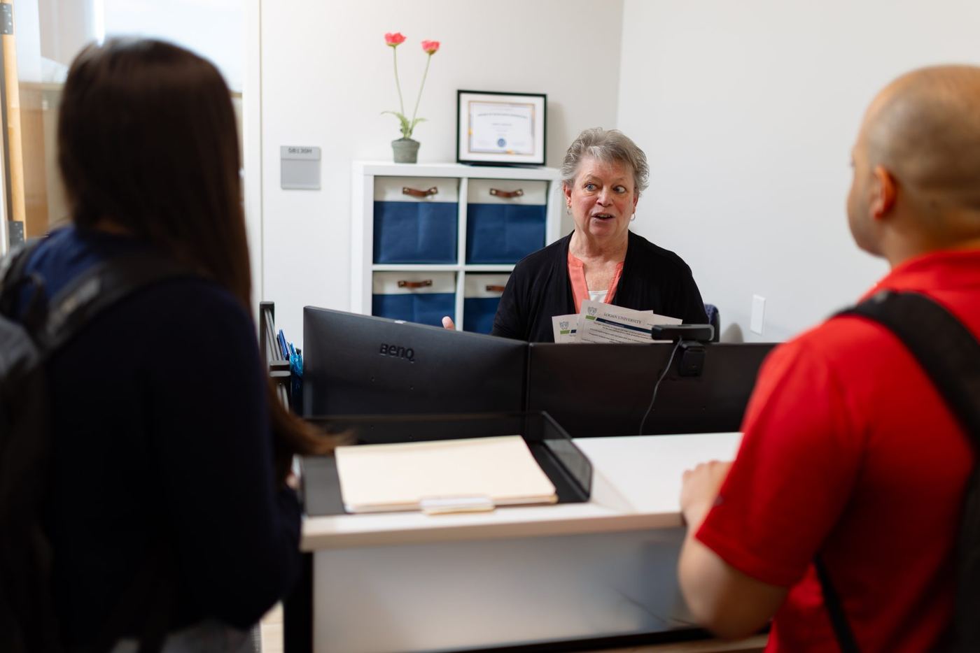 An older woman speaks with two students, one male and one female, at a reception desk in a bright office.