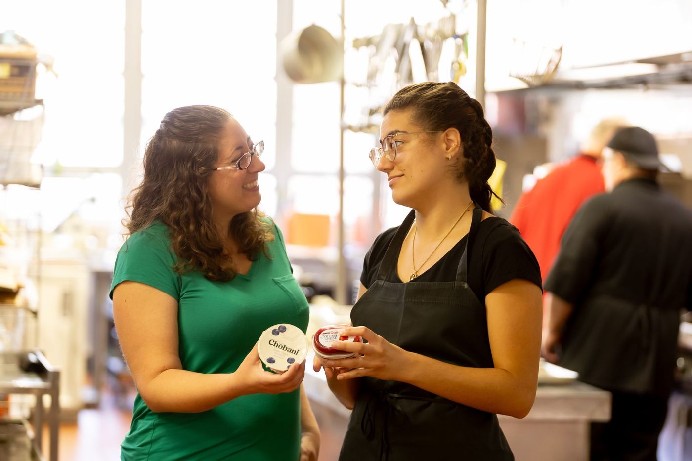 Two women, one in a green shirt and one in an apron, smile while holding yogurt and cottage cheese containers.