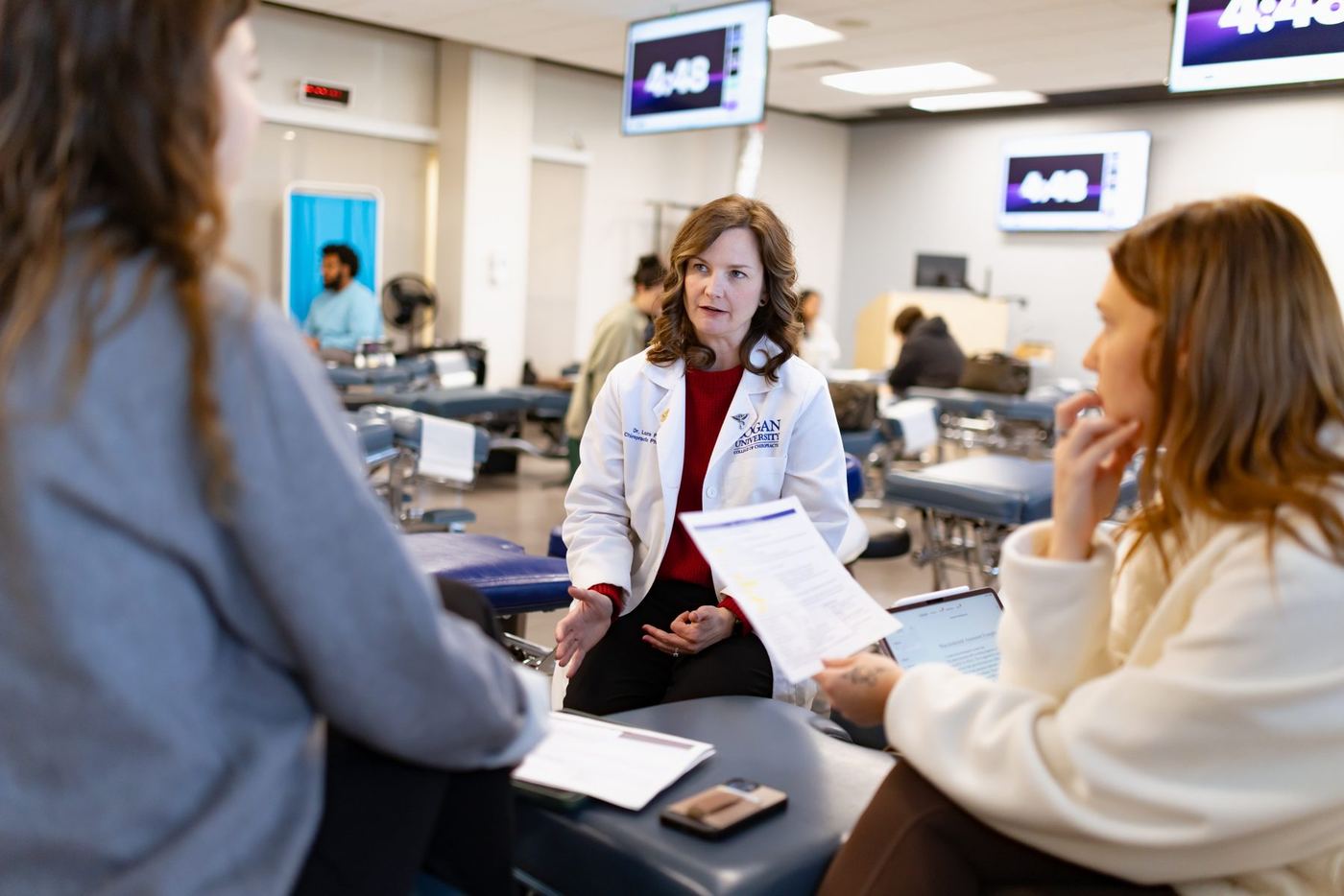 Dr. Laura P in a white lab coat talks with two female students in a bright classroom with exam tables.