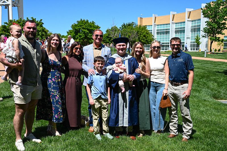 Logan graduate in regalia holding a baby, surrounded by family on the campus lawn at commencement