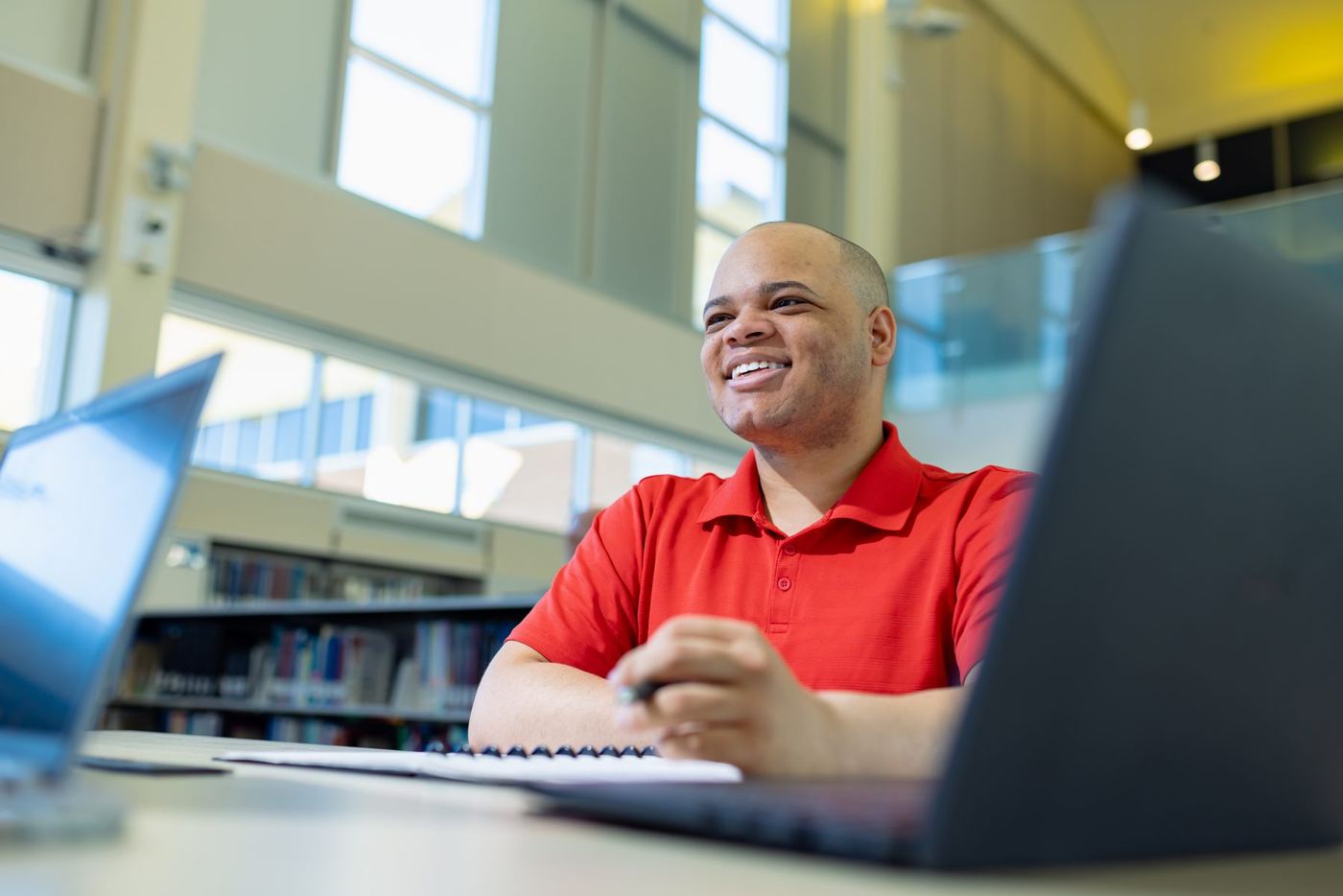Smiling student in red polo at a desk with two laptops and a notebook, engaged in study or conversation.