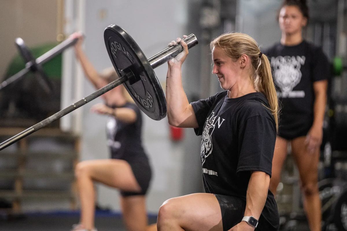 An athlete in a Logan Strength & Conditioning shirt presses a loaded barbell from a half-kneeling lunge