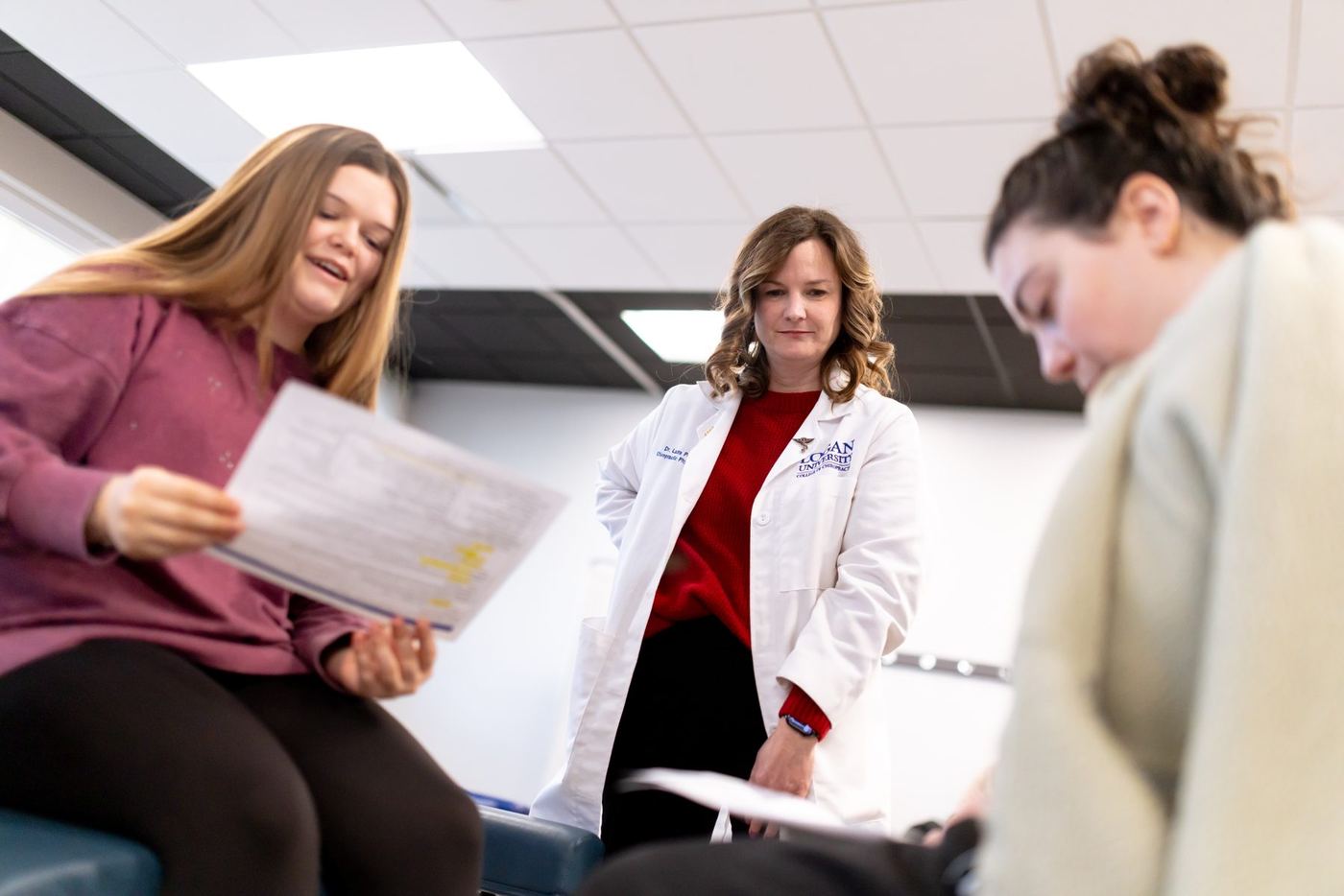 Doctor in lab coat stands between two young women, one smiling with a document, the other looking down.