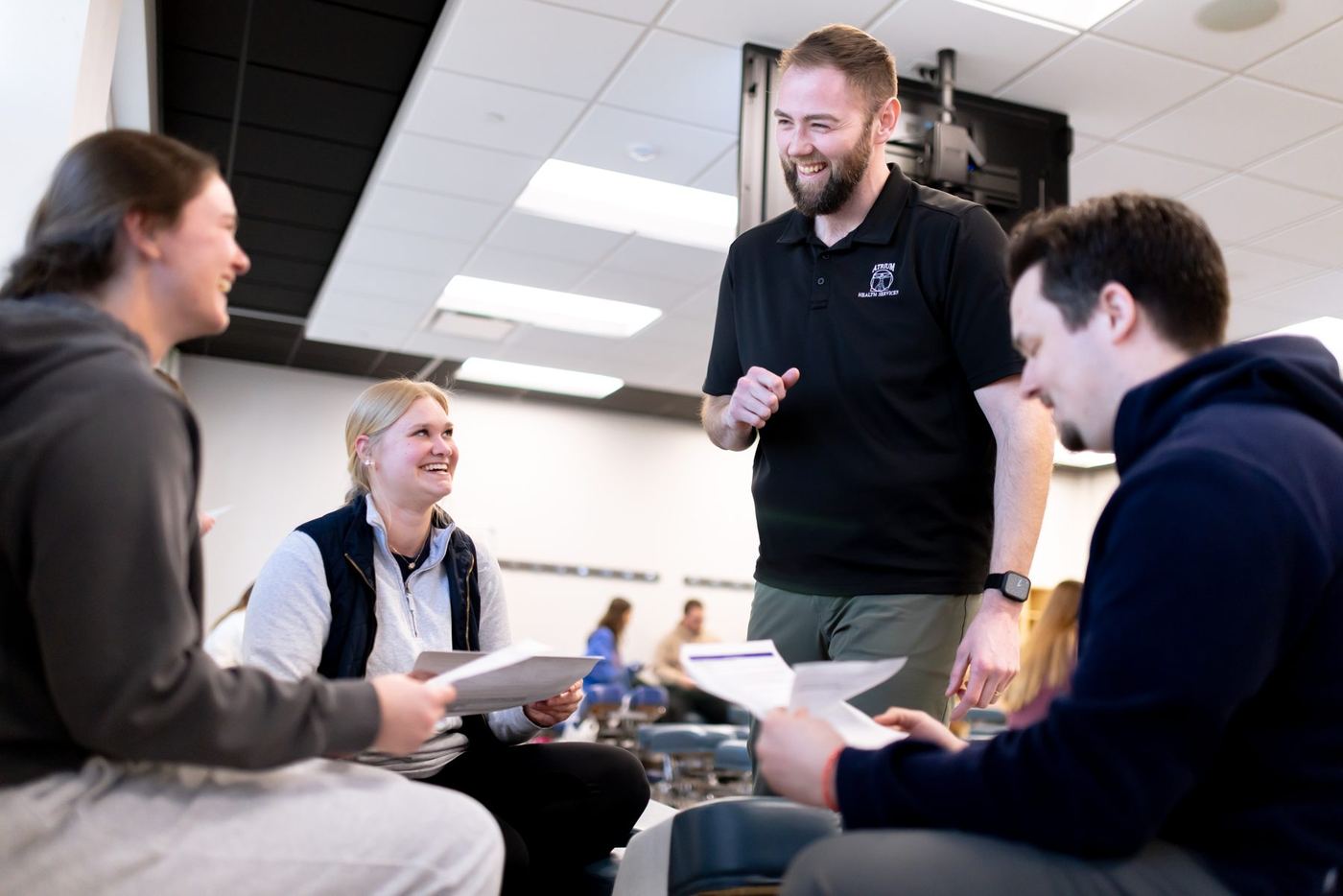 Smiling male instructor with Atrium Health logo interacts with three students holding papers in a bright classroom.