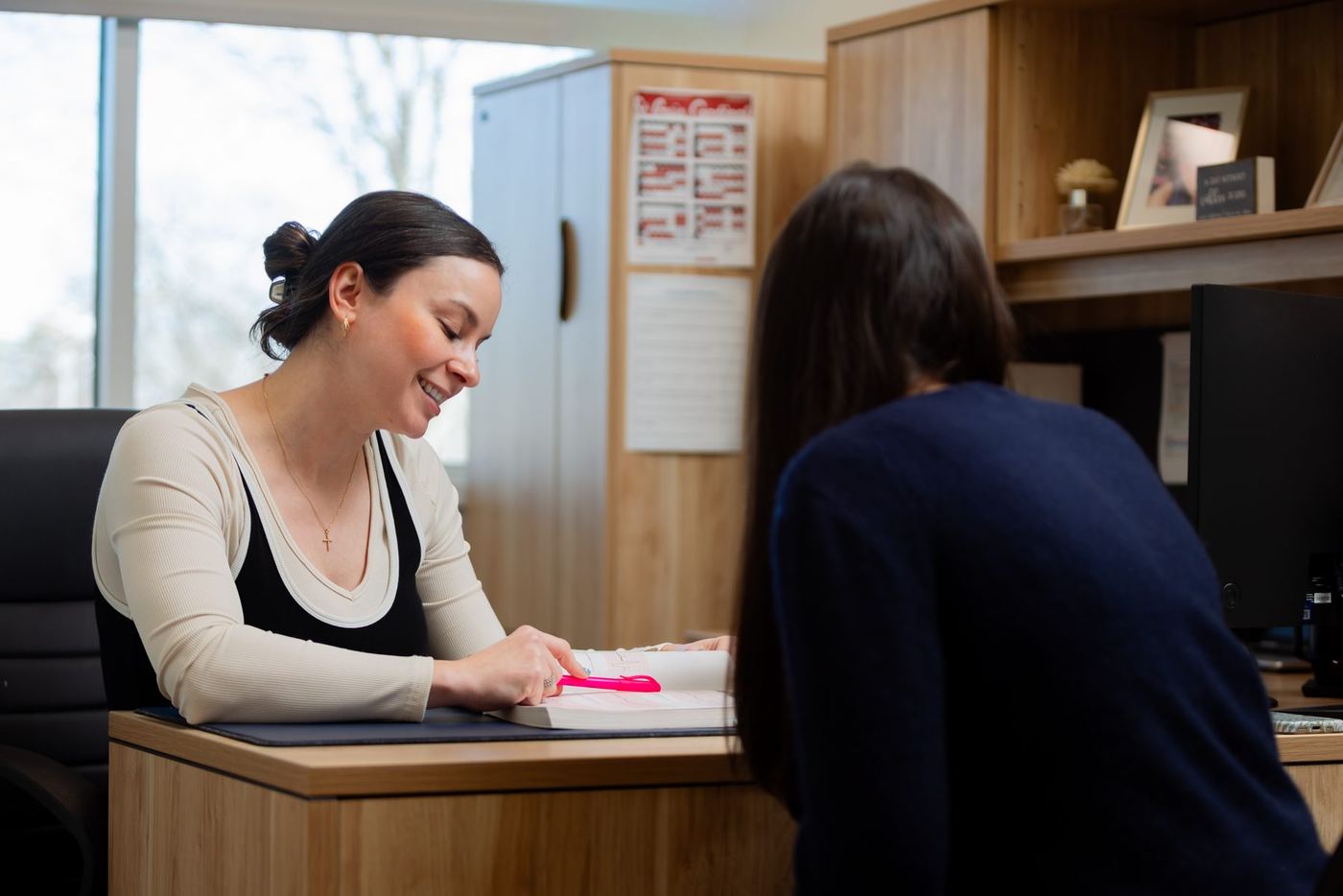 Smiling woman pointing at a book at a desk, with another person seated opposite her.