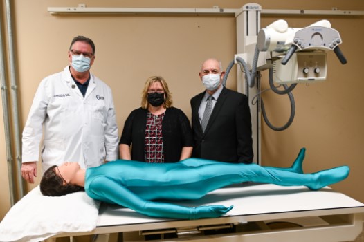 Three people stand behind a full-body radiology phantom lying on a table.
