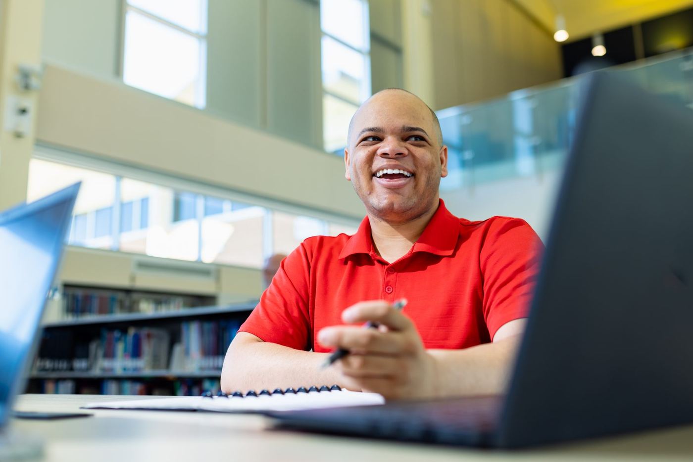 Bald man in red polo sits at desk with pen and notebook, smiling in a bright library space.