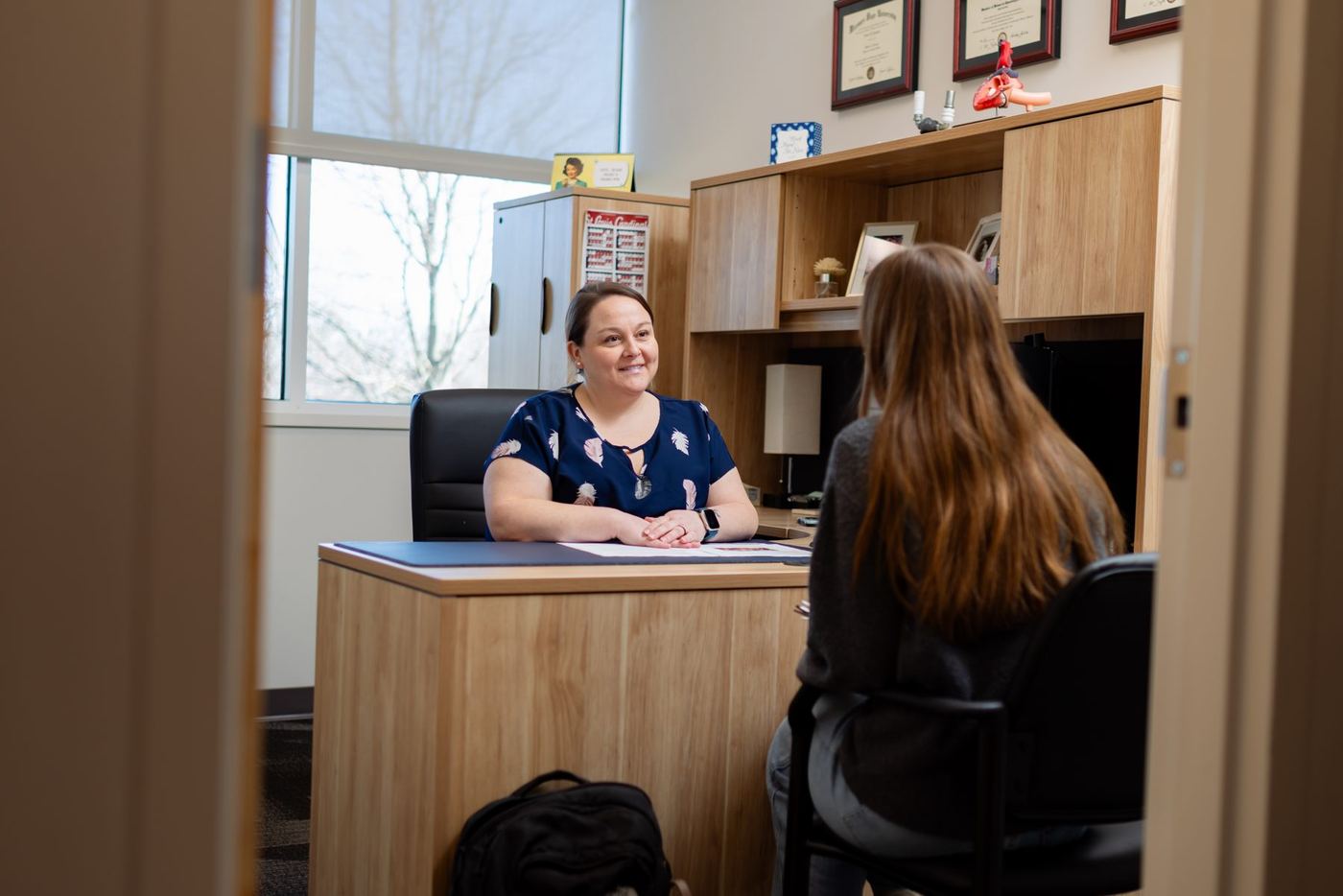 Smiling woman at desk talks to a student, with diplomas on shelves and a window in the background.