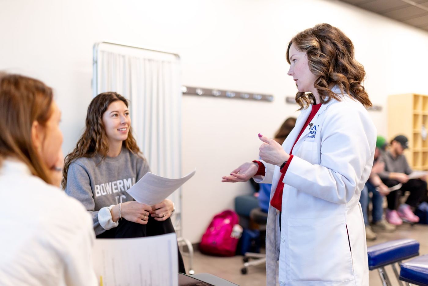 Faculty member in lab coat gestures while speaking to students in a classroom; one student smiles while listening.