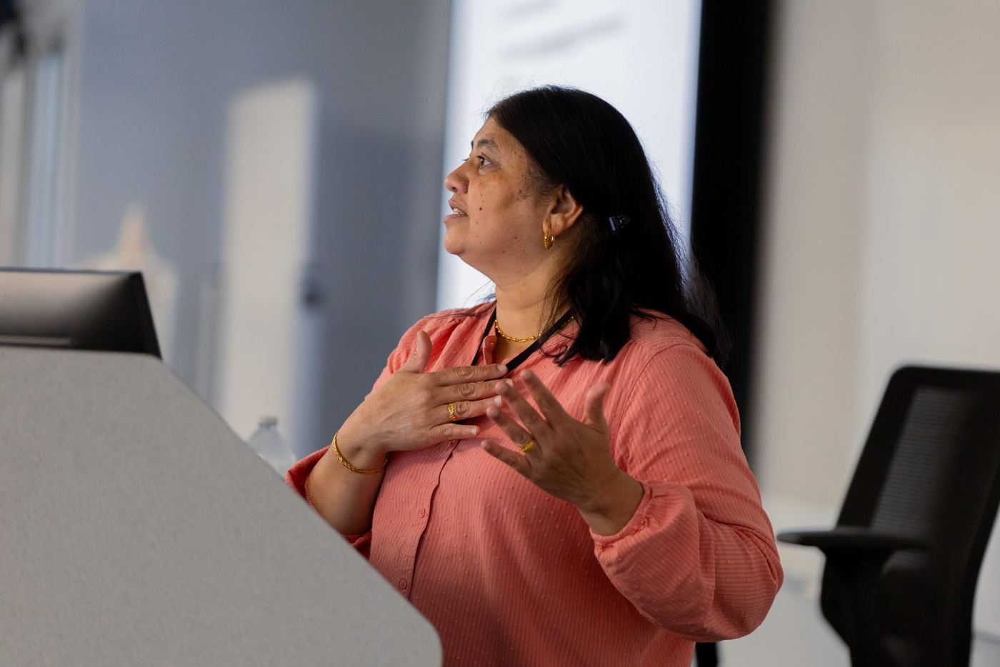 Woman in a coral shirt with gold jewelry gestures while speaking at a podium in a modern indoor setting.